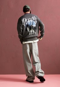 Szürke kapucnis pulóver "Death Valley" grafikával, fákkal és farkasokkal. Világosszürke, laza szabású nadrággal és fekete cipővel párosítva.