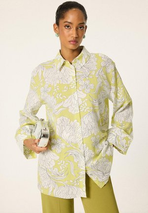 Femme portant une chemise jaune fleurie ample et un pantalon vert, tenant une petite pochette argentée d'une main.