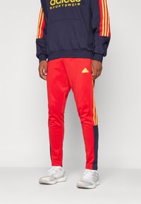 Pantaloni da tuta rossi con strisce gialle sui lati, caratterizzati da un accento blu navy. Realizzati in tessuto morbido, includono un piccolo dettaglio con logo.