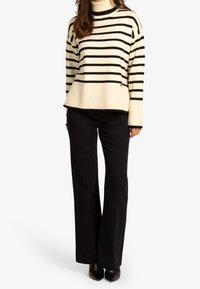 Pull à rayures crème et noir avec un col montant, un ourlet côtelé et une coupe décontractée, associé à un pantalon noir évasé et des chaussures noires à bouts pointus.