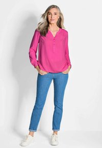 Fuchsia Bluse mit V-Ausschnitt und Knopfdetail, kombiniert mit hochgekrempelten hellblauen Jeans und weißen Sneakern. Glatter Stoff mit lockerer Passform.