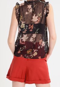 Gennemsigtig sort bluse med blomster mønstre, flæser på ærmerne og en hjerteformet åbning bagpå, parret med røde shorts med opkrøllede kanter.