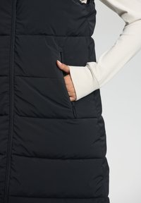Gilet rembourré noir avec une poche zippée et une texture lisse. Le gilet présente des coutures verticales et est stratifié pour l'isolation.