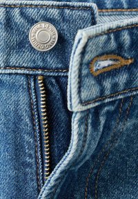 Gros plan d'un jean en denim bleu montrant un bouton en métal avec l'inscription "MNG DENIM" et une fermeture éclair en laiton partiellement ouverte avec des coutures orange.