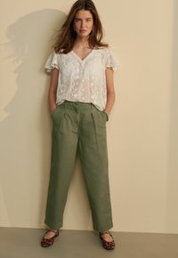 Witte kanten blouse met korte gerimpelde mouwen, olijfgroene high-waisted broek en flats met luipaardprint. Gladde texturen en een relaxte pasvorm.
