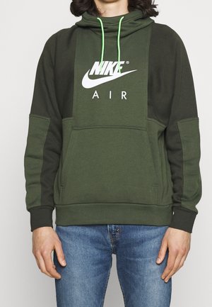 Person trägt einen zweifarbigen grünen Nike Air Hoodie mit neongrünen Kordeln und blauer Jeans, steht vor einem schlichten hellen Hintergrund.