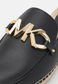Mocassino in pelle nera con un prominente logo MK in tono oro, caratterizzato da una finitura testurizzata e dettagli cuciti lungo il bordo.