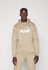Beige Kapuzenpullover aus weichem Material, mit einer Vordertasche, Kordelzugkapuze und weißem PUMA-Logo auf der Brust und dem Ärmel.