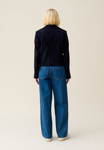 Veste en daim bleu marine au design ajusté, avec épaulettes, associée à un jean en denim bleu ample et des baskets blanches.