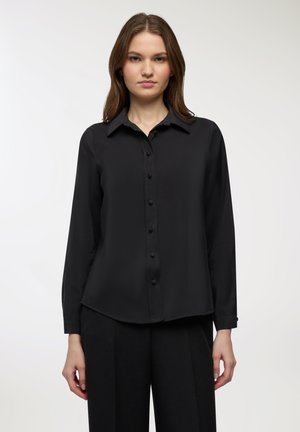 Giovane donna che indossa una camicia nera a maniche lunghe con bottoni e pantaloni neri, in piedi contro uno sfondo grigio chiaro.