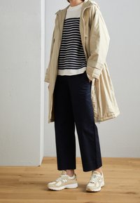 Cappotto lungo beige, maglione a righe blu navy, pantaloni larghi blu navy scuri e sneakers crema con accenti chiari. Look casual, vestibilità rilassata e look a strati.