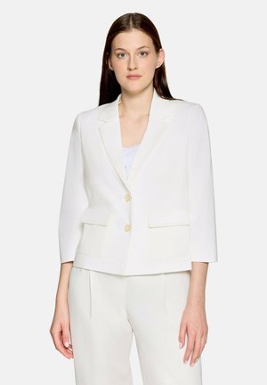 Kobieta o długich brązowych włosach, ubrana w biały dopasowany żakiet z złotymi guzikami i dopasowane białe spodnie na jednolitym tle.