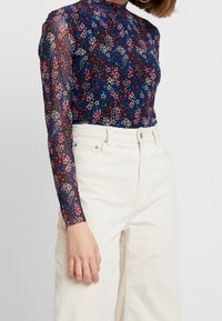 Vrouw die een doorschijnende navy blouse met lange mouwen en bloemenprint draagt, ingestopt in een hoge taille crème kleurige corduroy broek, staand tegen een effen lichte achtergrond.