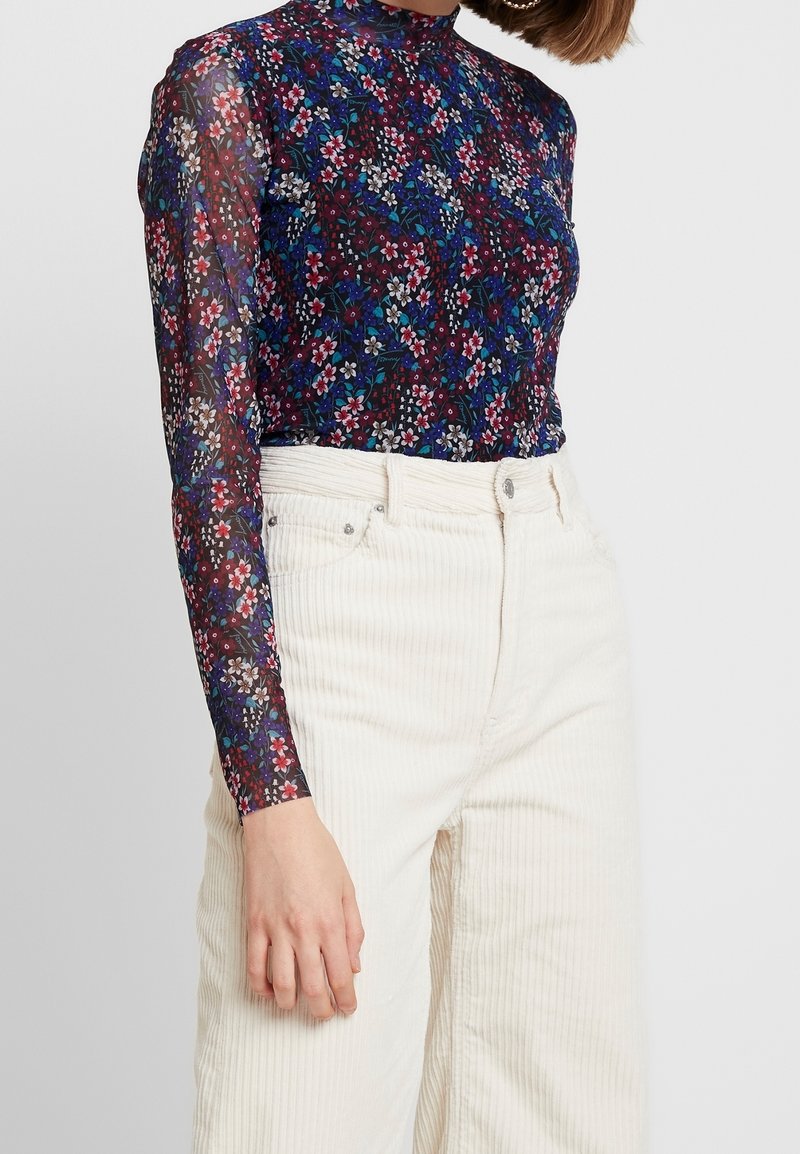 Vrouw die een doorschijnende navy blouse met lange mouwen en bloemenprint draagt, ingestopt in een hoge taille crème kleurige corduroy broek, staand tegen een effen lichte achtergrond.