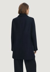 Cappotto blu navy con un design strutturato, maniche lunghe e una singola fessura posteriore. Realizzato in lana con una texture liscia. Aspetto minimalista senza hardware visibile.