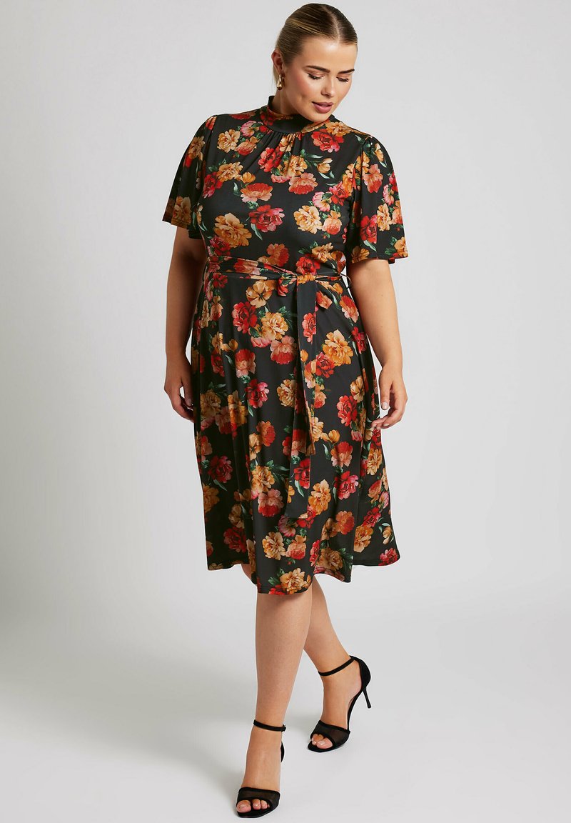 Vestido floral negro con mangas cortas, adornado con flores rojas y naranjas, con un cinturón en la cintura ceñida y una longitud midi fluida. Sandalias de tacón alto negras.