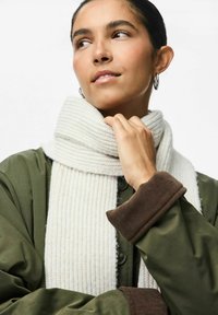 Écharpe blanche à côtes drapée autour du col d'une veste vert foncé avec des poignets en velours côtelé marron, mettant en valeur une texture de tricot douce et des tons neutres.