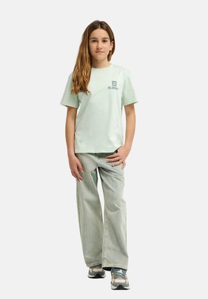 Jasnozielona bawełniana koszulka z krótkimi rękawami i logo na piersi, zestawiona z szerokimi dżinsami w jasnym odcieniu i wielokolorowymi sneakersami.
