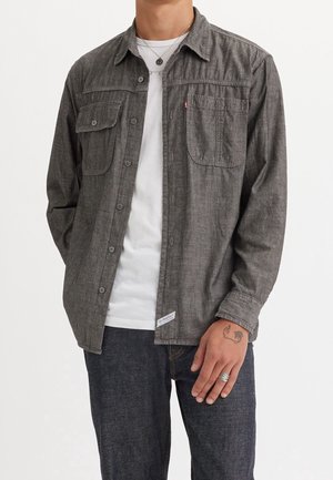 Mann trägt ein aufgeknöpftes graues Langarmhemd über einem weißen T-Shirt, dunkle Jeans, sichtbare Handtattoo und einen silbernen Ring am Finger.