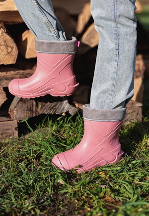 Rosa Gummistiefel mit strukturierter Oberfläche und grauen Strickbündchen, mit einem markanten Design und profilierten Sohlen, vor Gras und Holzstämmen platziert.