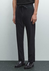 Personne portant un pantalon noir ajusté avec taille élastique et des mocassins en cuir noirs, se tenant sur un sol en terrazzo sombre.