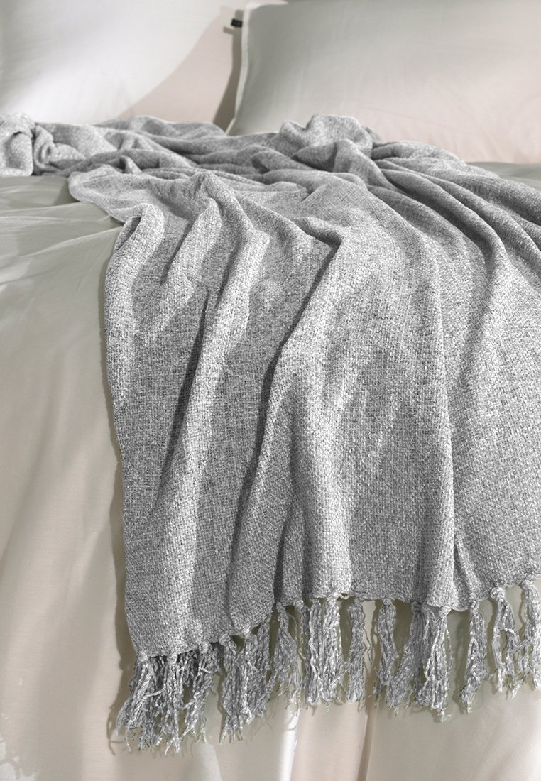 Šedý tkaný přehoz s texturovaným povrchem a třásněmi, navrstvený na světle zbarveném prostěradle, vytváří měkký a lákavý vzhled.