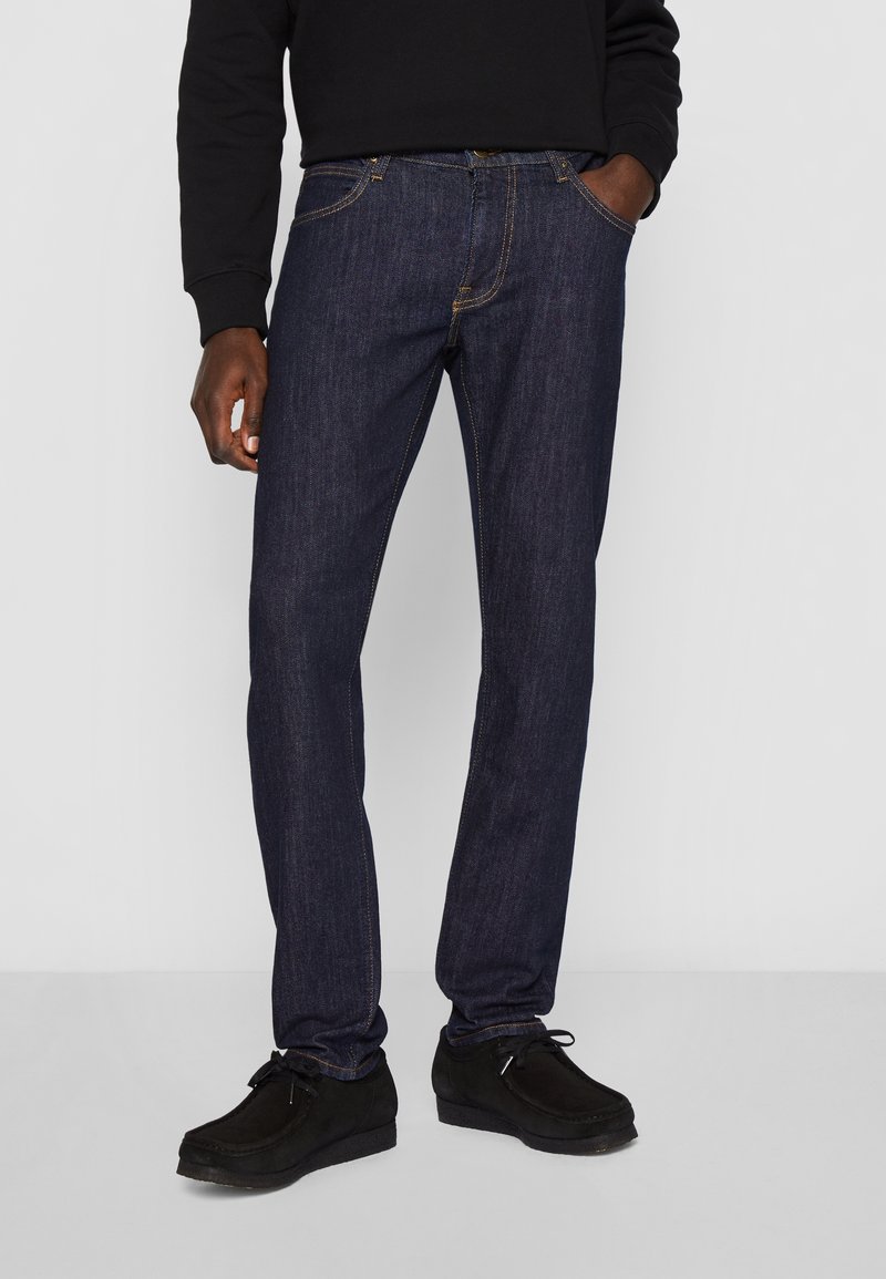 Homme portant un jean droit bleu foncé, des chaussures noires à lacets et un haut noir à manches longues, une main dans la poche.