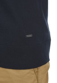 Marineblauer Strickpullover mit geripptem Saum und einem kleinen Metalltag mit der Aufschrift "JEANSCODE" nahe dem unteren Ende. Getragen mit khakifarbenen Hosen.