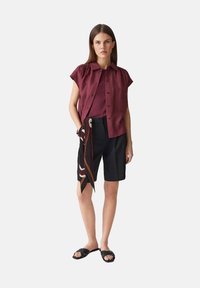 Camicia corta bordeaux sopra un top bordeaux, abbinata a pantaloni corti neri e una sciarpa a fantasia annodata in vita. Sandali a scivolo neri.