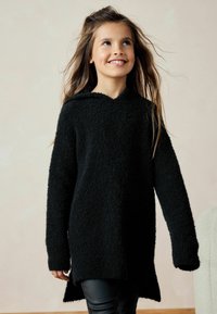 Sweat à capuche noir avec une finition texturée et duveteuse, présentant de longues manches et un ourlet arrière plus long, porté avec un pantalon noir.
