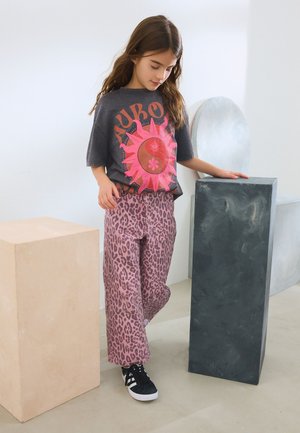 Camiseta gráfica gris con diseño de sol rosa combinada con pantalones de leopardo rosa. Zapatillas negras. Fondo minimalista con formas geométricas.