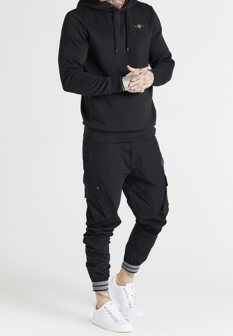Homme portant un sweat à capuche noir et un pantalon cargo jogger noir avec des revers roulés et des baskets blanches, debout les mains jointes.