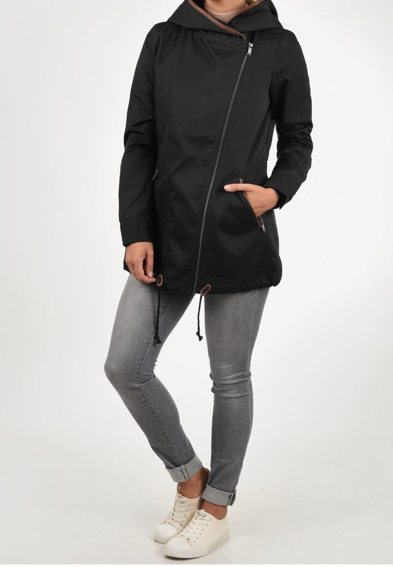 Black hooded jacket with a diagonal zipper, two side pockets, and adjustable drawstring hem, paired with gray jeans and white sneakers.