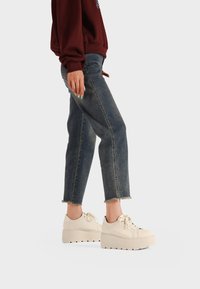 Sneakers bianchi con suola spessa, lacci e dettagli testurizzati, abbinati a jeans in denim blu scuro con orli sfrangiati e un fit rilassato.