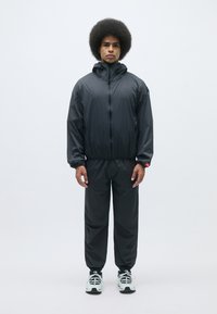 Survêtement noir léger comprenant une veste à capuche zippée et un pantalon assorti. Il possède des poignets et une taille élastiques ; tissu lisse et résistant à l'eau.