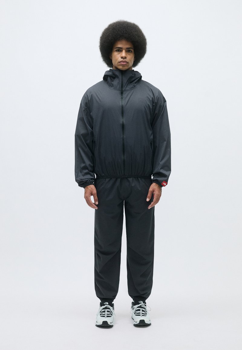 Survêtement noir léger comprenant une veste à capuche zippée et un pantalon assorti. Il possède des poignets et une taille élastiques ; tissu lisse et résistant à l'eau.