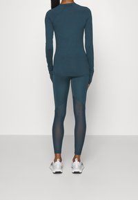 Töjbar, långärmad topp i teal och matchande leggings med meshdetaljer; båda är åtsittande och tillverkade av stretchmaterial, ihop med vita träningsskor.