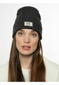Bonnet noir tricoté avec un motif texturé et une étiquette rectangulaire beige portant l'inscription "NXG PROTEST". Matériel doux et extensible qui s'ajuste parfaitement.