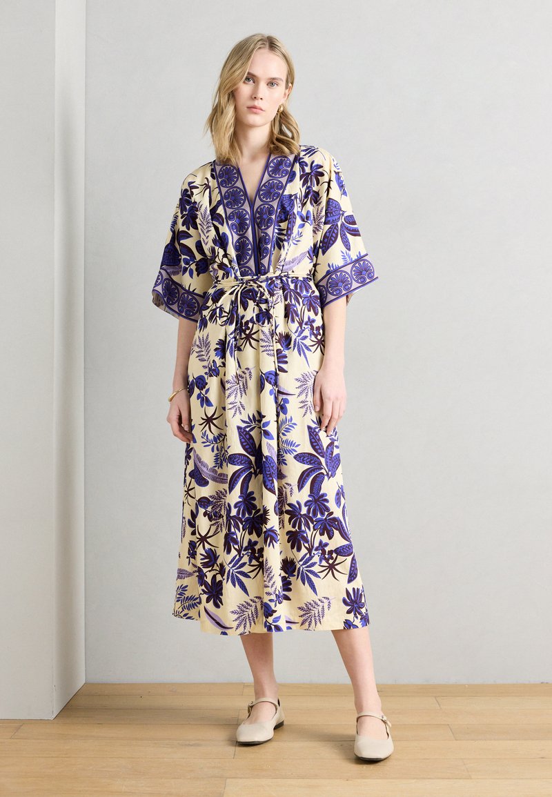 Γυναίκα στέκεται σε εσωτερικό χώρο φορώντας ένα μπεζ φόρεμα με μπλε λουλουδένια σχέδια, μανίκια μέχρι τον αγκώνα και ζώνη στη μέση, συνδυασμένο με μπεζ παπούτσια.