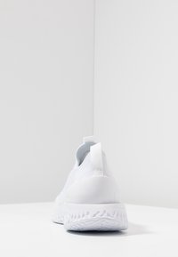 Weiße Sportschuhe mit strukturiertem Strickobermaterial, geformter Ferse und Gummisohle mit Wellenmuster. Verfügt über eine Lasche zum einfachen An- und Ausziehen.