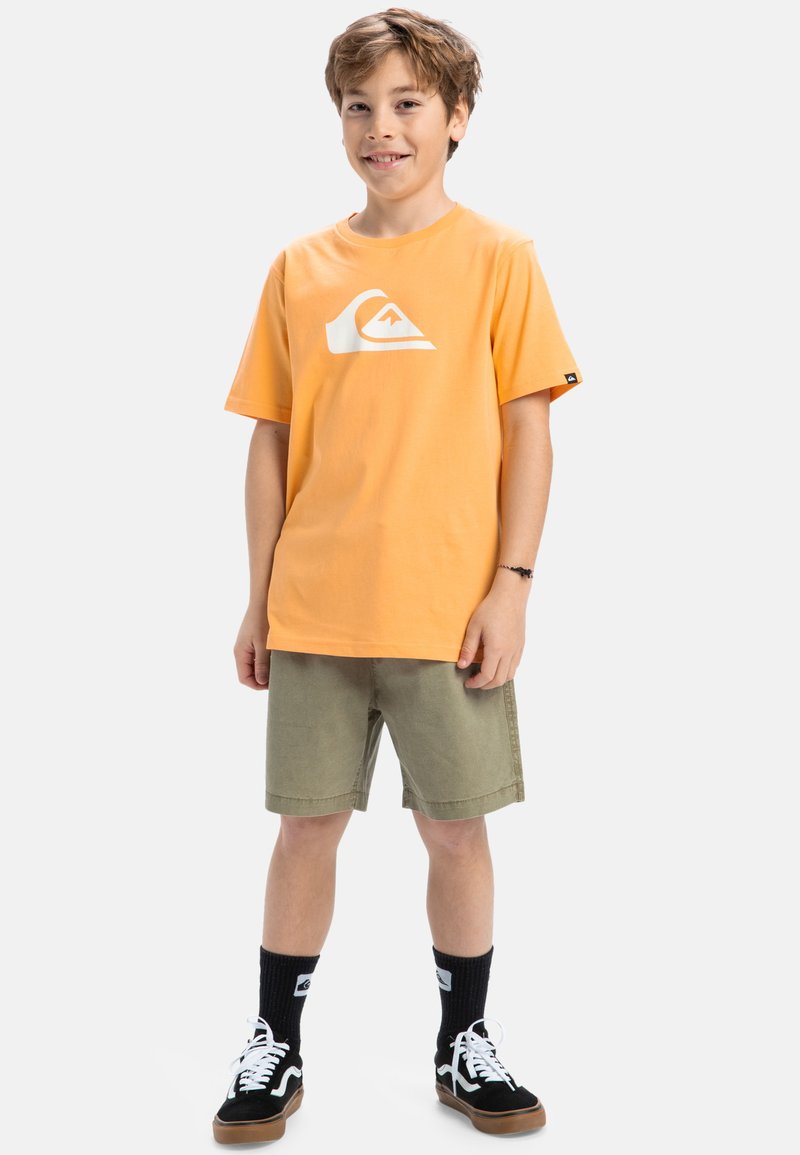 Camiseta de algodón naranja con logo blanco, combinada con pantalones cortos verde oliva, zapatillas en blanco y negro, y calcetas negras hasta la rodilla.