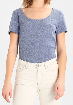 Femme portant un t-shirt bleu ajusté à manches courtes et col rond, rentré dans un jean taille haute blanc cassé avec poches avant et fermeture par boutons.