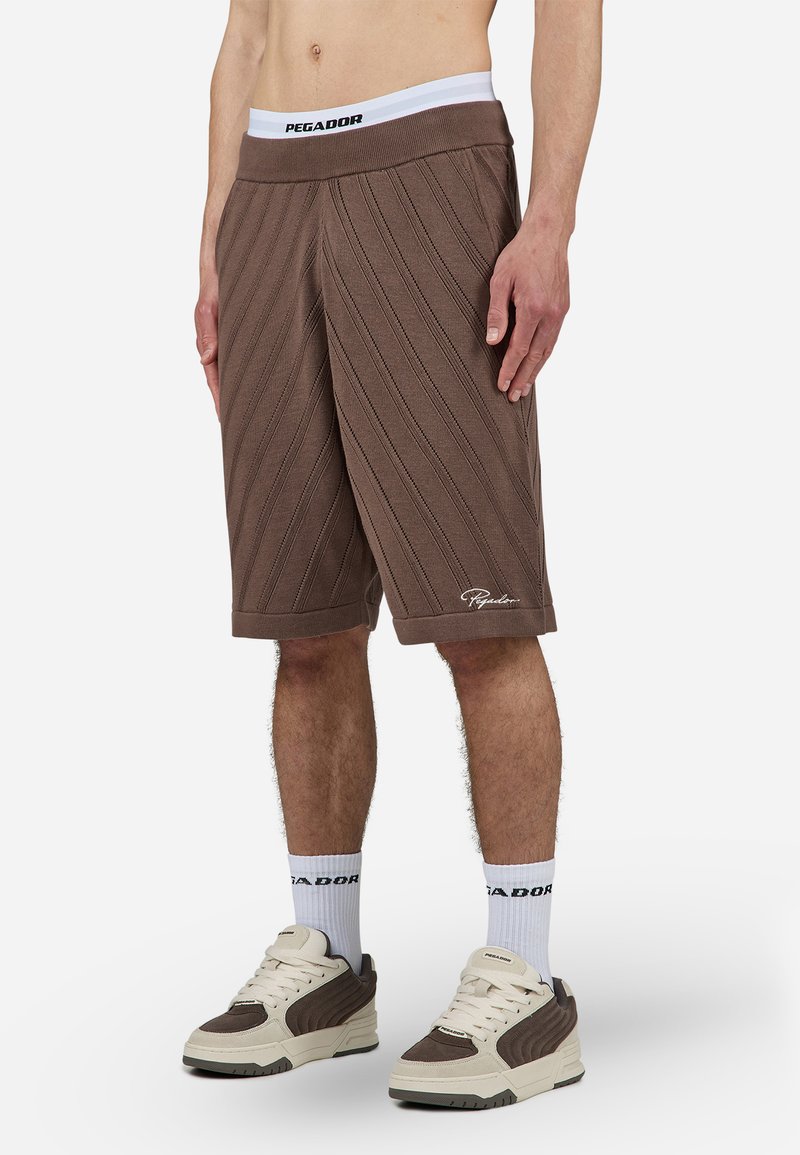 Brune strikkede shorts med diagonalt ribbestrikket tekstur, elastisk midje som har "PEGADOR," og en hvit logo ved kanten.