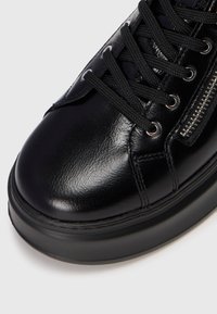 Close-up of black leather sneaker with thick sole, black laces, and side zipper detail on white background.