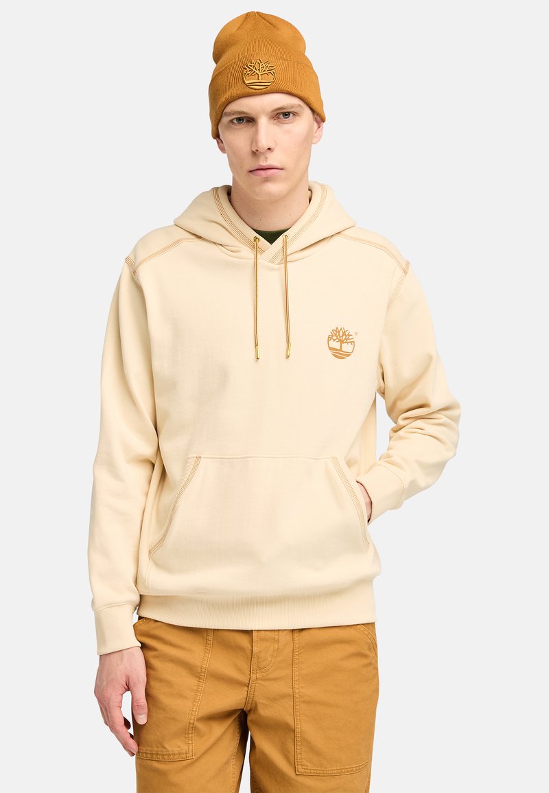 Sudadera color crema con un bolsillo delantero y detalles en dorado, combinada con un gorro mostaza a juego que lleva un logotipo bordado.