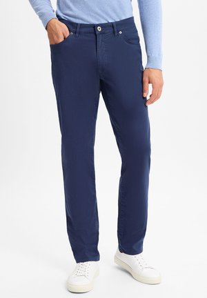 Marineblaue Hose aus weichem Stoff, mit geradem Schnitt, zwei seitlichen Taschen und einem Knopf mit Reißverschluss. Getragen mit weißen Turnschuhen.
