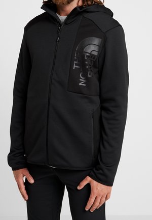 Mand iført sort lynlåshættejakke med stort sort på sort The North Face-logo på venstre brystlomme.