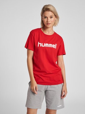 Femme portant un t-shirt rouge "hummel" et un short gris "hummel", debout devant un fond gris clair uni.