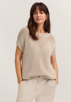 Woman with long brown hair wearing gold hoop earrings, a loose beige knit short-sleeve top, and light beige pants with hands in pockets.