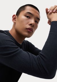 Dunkelblaues Sweatshirt aus weichem, strukturiertem Strick mit Rundhalsausschnitt. Nahaufnahme eines männlichen Modells, das Arm- und Gesichtsdetails präsentiert.